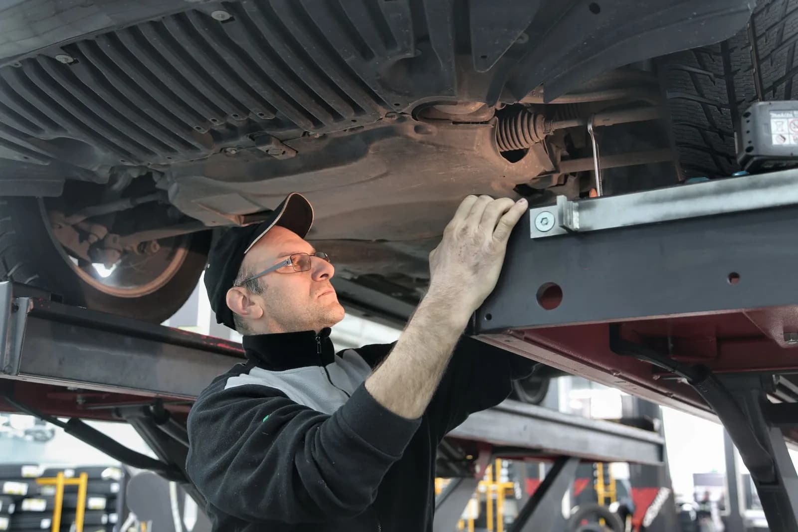 Mann mit Brille arbeitet stehend am Unterboden eines Autos auf der Hebebühne